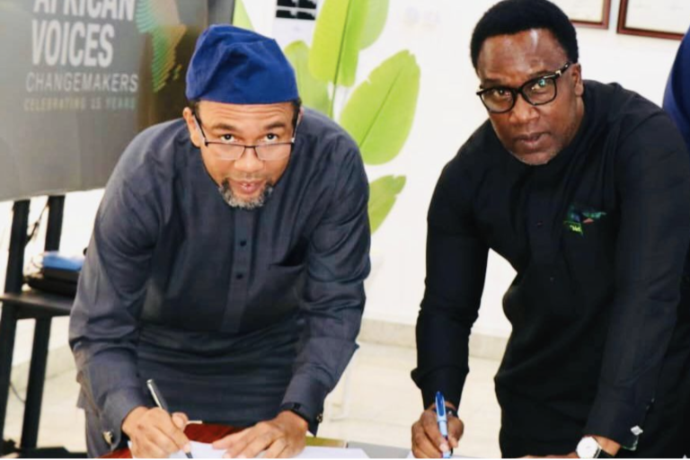 Two men in dark clothing are leaning over a table, holding pens, and writing on documents. A sign partially reads "African Voices Changemakers" in the background. .