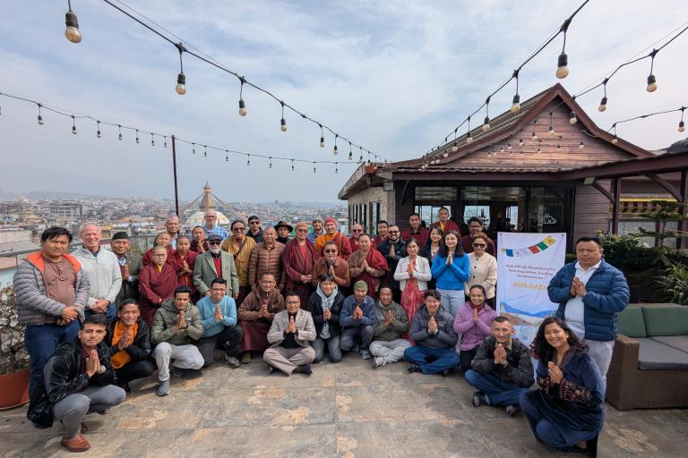 A group of people pose for a photo on a rooftop with string lights, a city view, and a banner that reads "HIA Daily." Most of them have their palms together in a greeting gesture.