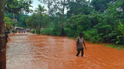 A person walks through a flooded, muddy street surrounded by dense green vegetation and trees, with other people visible in the background.