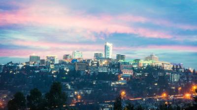 A brightly lit cityscape at dusk with a prominent modern building in the center, surrounded by smaller buildings under a sky with pink and blue hues.