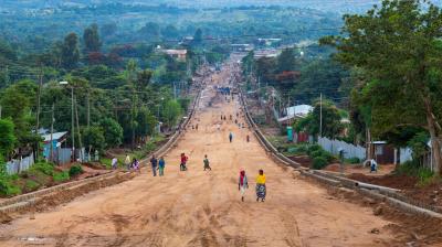 A wide, unpaved road with people walking and greenery on both sides, stretching into the distance.