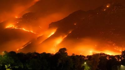 A wildfire engulfs a mountainside at night, with intense orange flames and smoke illuminating the scene. Trees and a few lights are visible in the foreground.