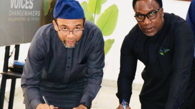 Two men in dark clothing are leaning over a table, holding pens, and writing on documents. A sign partially reads "African Voices Changemakers" in the background. .