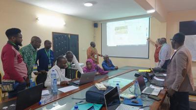 People engaged in a meeting, some seated at a long table with laptops, while one person presents information on a projected screen.