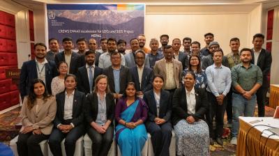 A group of men and women in business attire pose for a photo in a conference room with a banner reading "CREWS EW4All Accelerator for LDCs and SIDS Project Nepal" in the background.