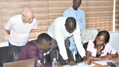 Five people are gathered around a table with papers, a laptop, and water bottles, engaged in a discussion and reviewing documents in an office setting.