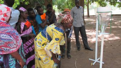 A group of people observes a demonstration of a water collection device outdoors under trees, with a woman in the foreground leaning in for a closer look.