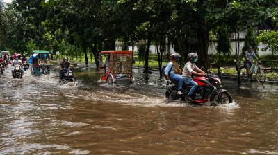 Motorcycles, rickshaws, and bicycles travel through a heavily flooded street lined with trees during daytime.