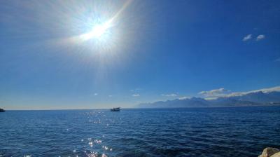 The sun shines brightly over a calm blue sea with a boat in the distance and mountains visible on the horizon under a clear sky.