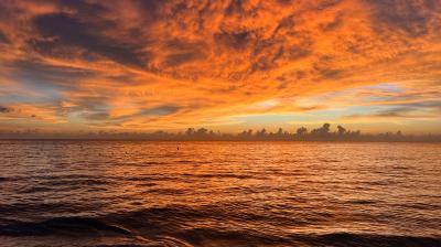 Orange and gold clouds fill the sky over a calm ocean at sunset, reflecting warm colors on the water and gentle waves near the shore.