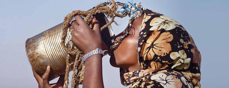 Person drinking from a large, weathered tin can against a clear sky, wearing floral patterned clothing and a wristwatch.