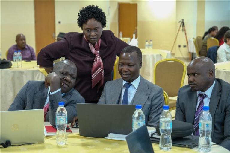 Four people in business attire sit and stand around a table with laptops and water bottles, appearing to discuss something during a meeting or workshop.